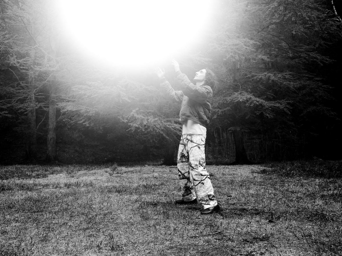 Little Yarmouth EP. 2. A figure reaching toward bright light in a monochrome forest clearing.