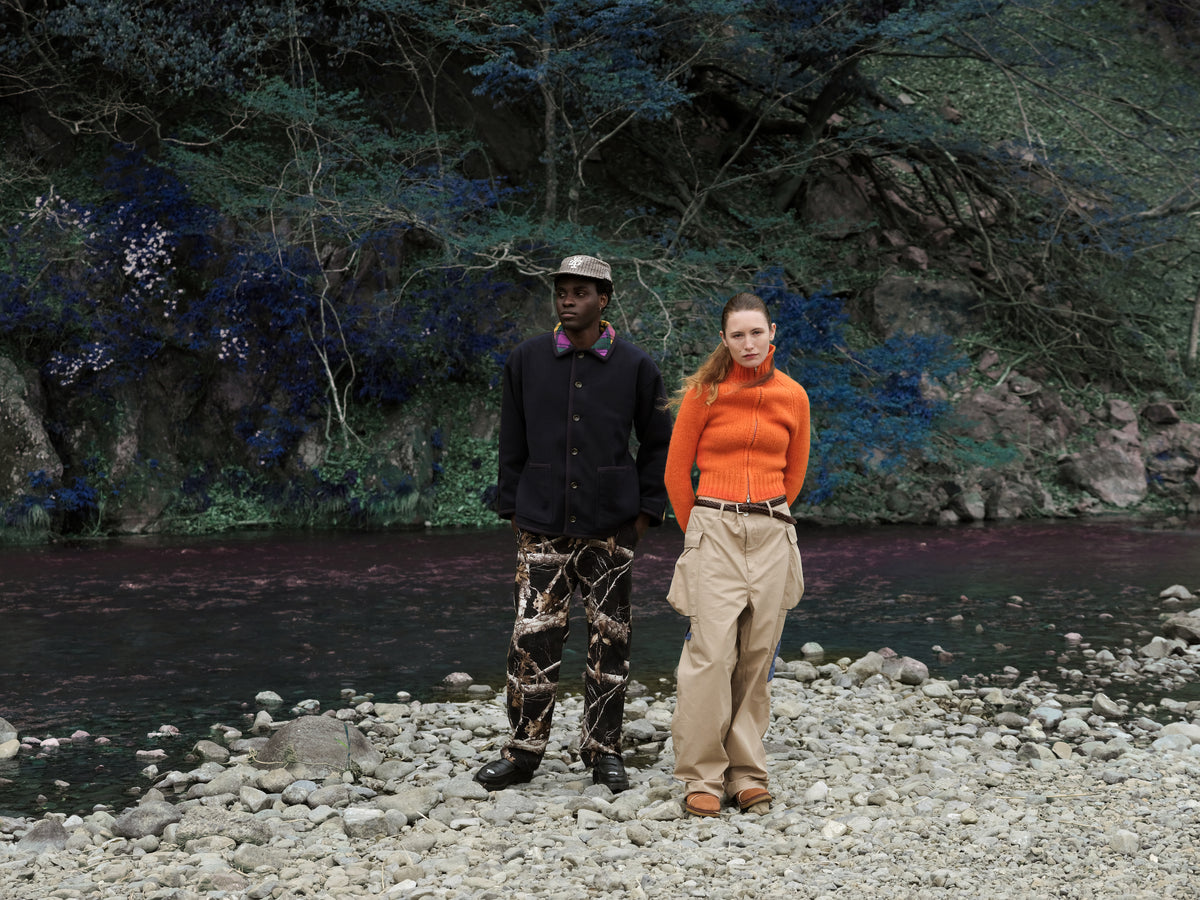 Little Yarmouth EP. 2. Two figures in a dark jacket and orange knit stand on a rocky riverbank.